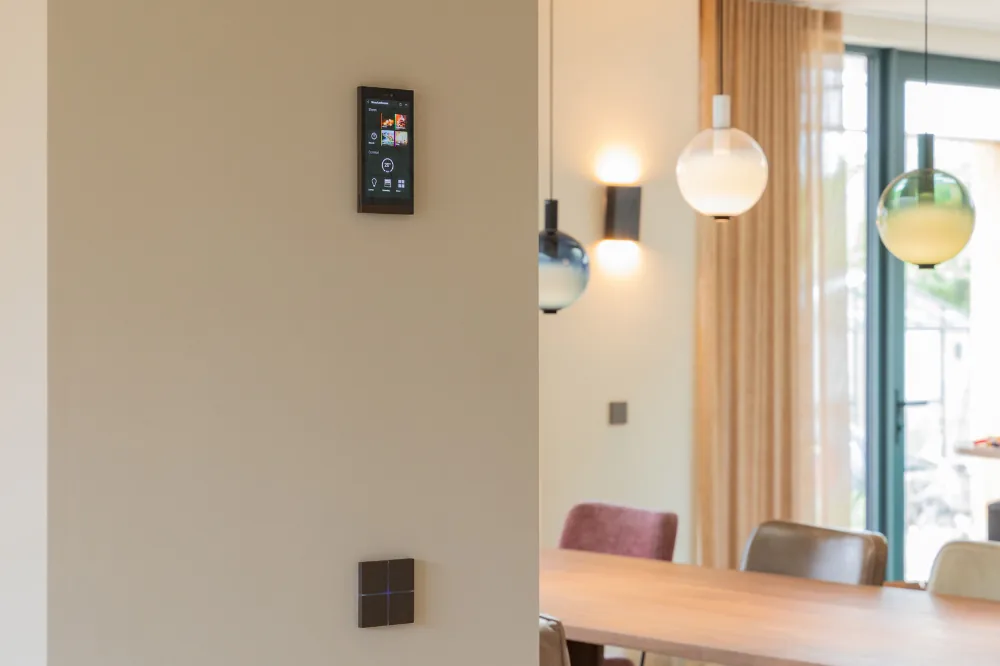 Basalte Ellie touchscreen en Sentido schakelaar aan een wand, met zicht op design hanglampen en een eethoek